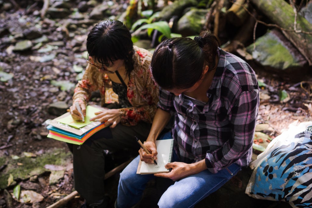 Two people sit in the jungle bent over notebooks and colored paper, one with a pen and one with a marker. 