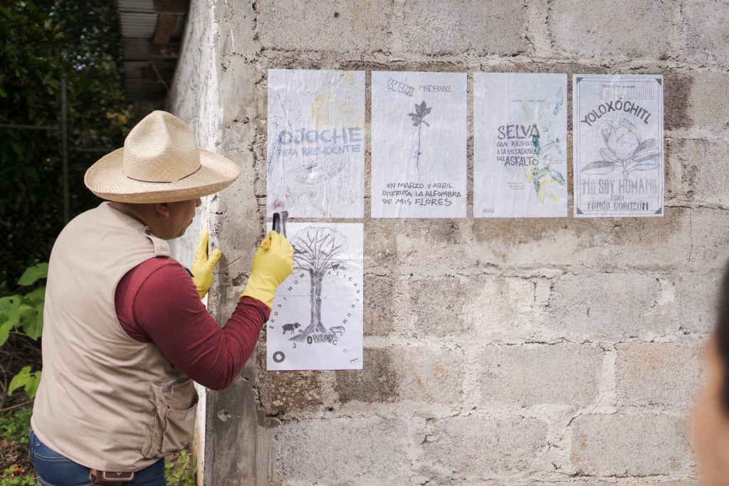 A person in a brimmed hat pastes five posters to a cement brick wall. 