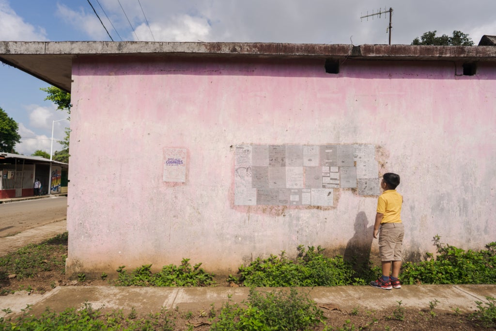 A boy in a yellow shirt an khaki pants looks up and to the left at a series of posters pasted in a rectangle on the pink wall of a concrete building.