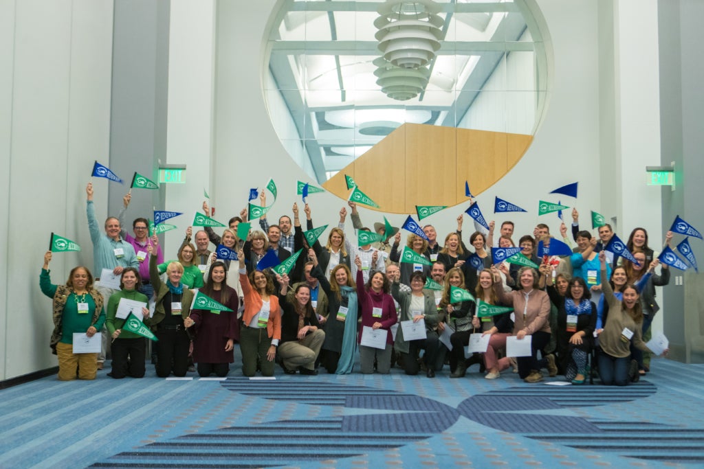 A group of newly accredited land trust leaders gather in celebration to wave blue and green accreditation flags while posing for a picture.