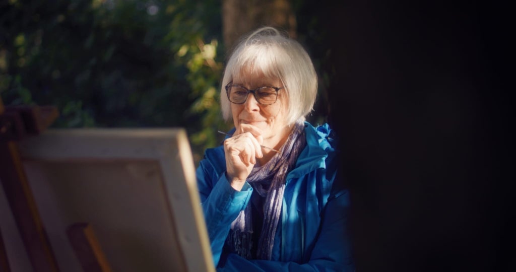 Sue Prince is dressed in a blue coat and lavender scarf. She holds one hand up to her face, a paintbrush in her grasp, as she considers the canvas before her.