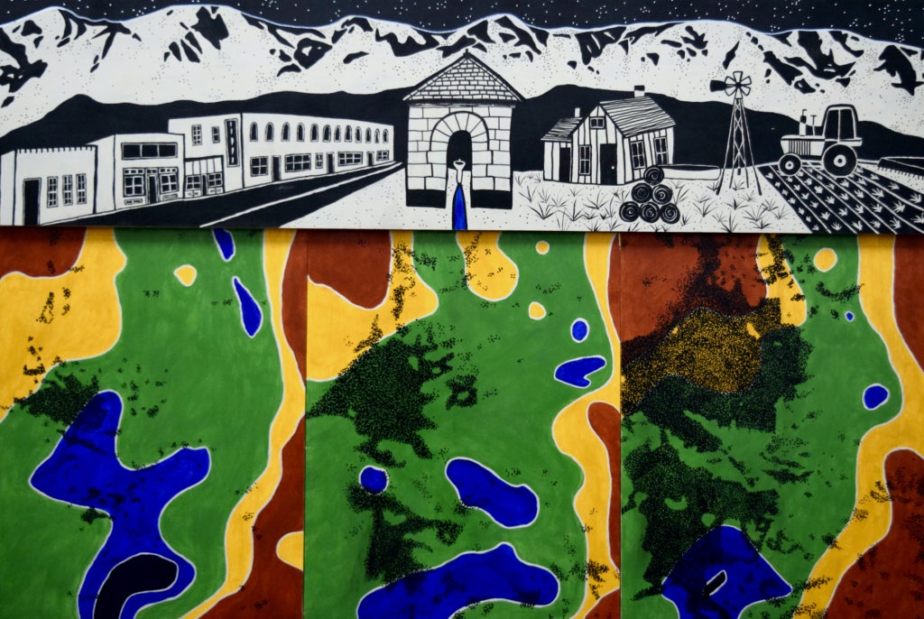 A black and white village at the foot of a mountain range is cut through by blue water and rests atop topographical heat maps in green, blue, yellow, and red. 