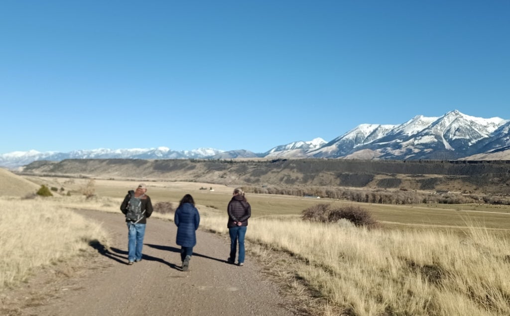 Three people walk down a dirt road in Montana, surrounded by grasslands that stretch toward snowcapped mountains.