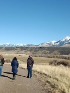 Three people walk down a dirt road in Montana with snow capped mountains in the background.