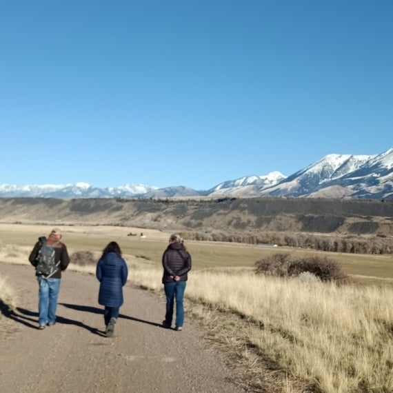 Three people walk down a dirt road in Montana with snow capped mountains in the background.
