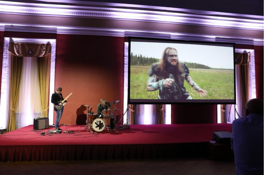 A guitarist and drummer play on a red stage, positioned to the far left. To their right is a large screen showing the image of a warrior in a field. 