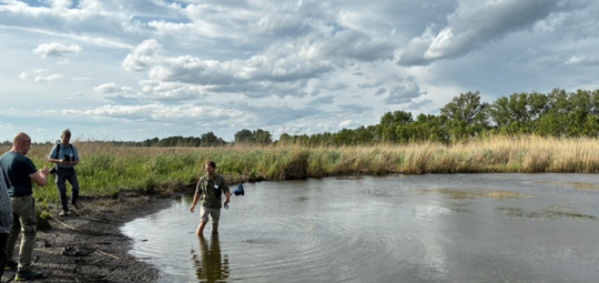 One man wades in a shallow natural pool while two others look on from the shore.