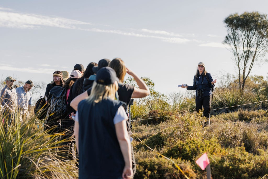 A woman speaks to a group of 10 in a scrubby landscape.