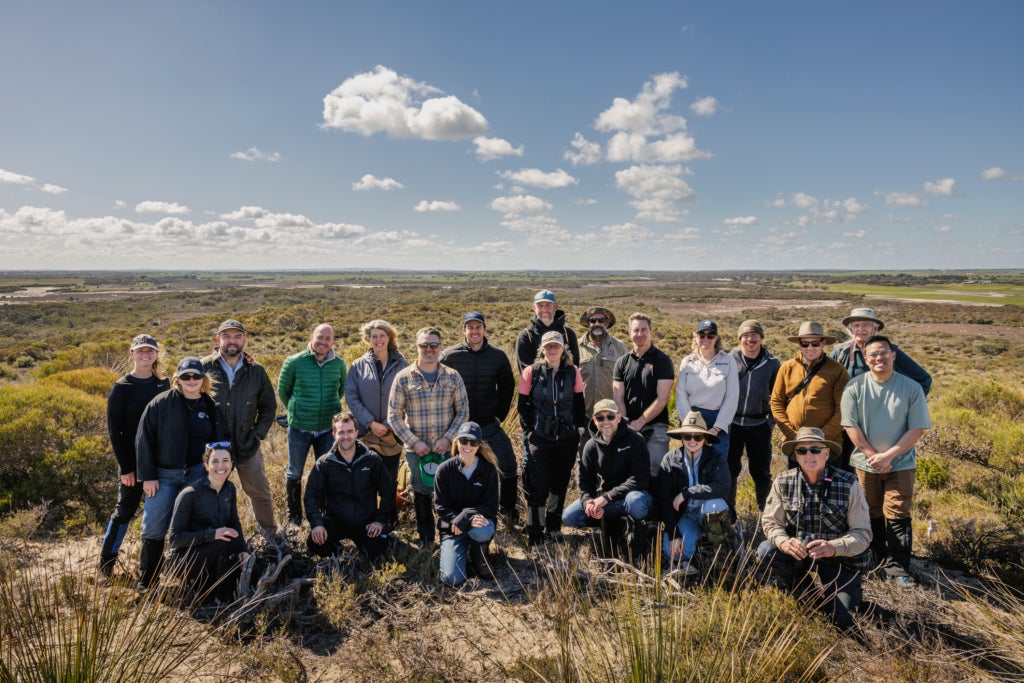 Two dozen people pose for a photo in a vast grassy landscape.
