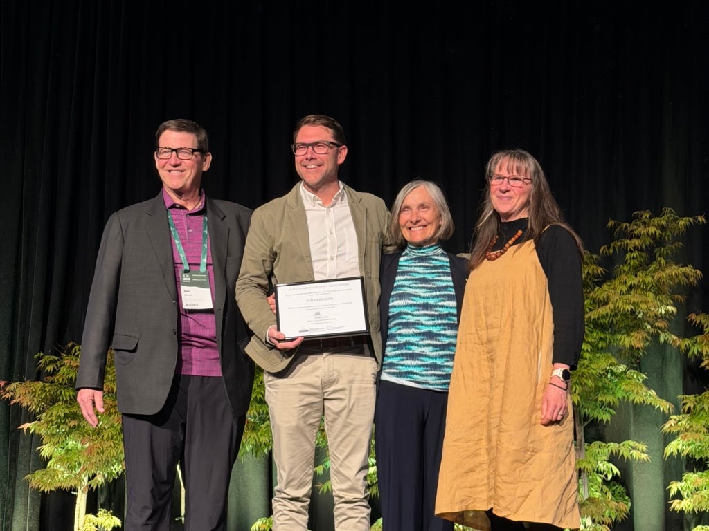 Four people smile on a conference room stage, with one holding an award certificate.